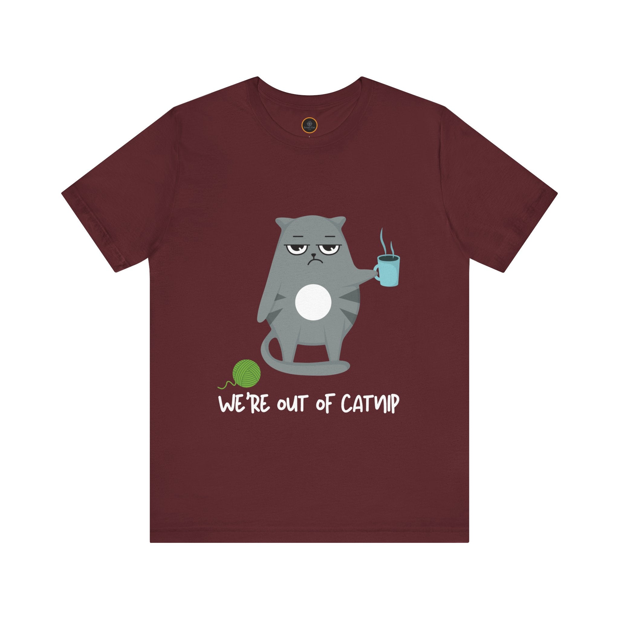 Crewneck t-shirt featuring a cartoon gray and white cat smugly holds a steaming cup of coffee. A green ball of yarn sits on the floor nearby. Below the image, the text reads "We're Out Of Catnip".