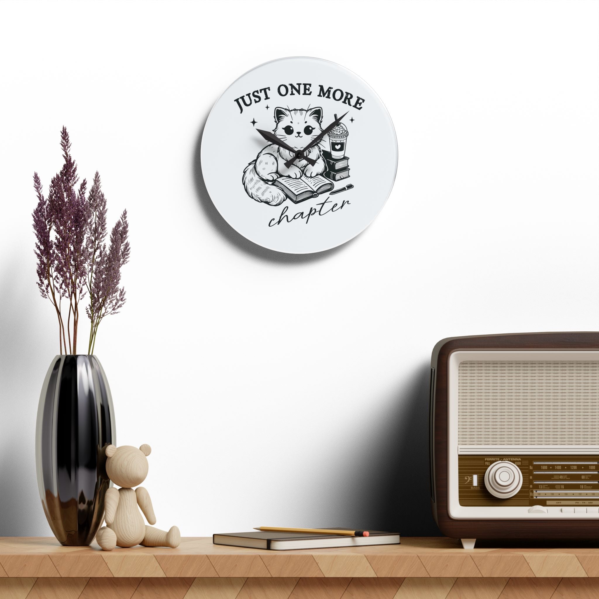Line art graphic on the "Just One More Chapter" Acrylic Wall Clock depicting a cat reading an open book beside a book stack topped with a beverage. The words "just one more" are above, and "chapter" in script is below.