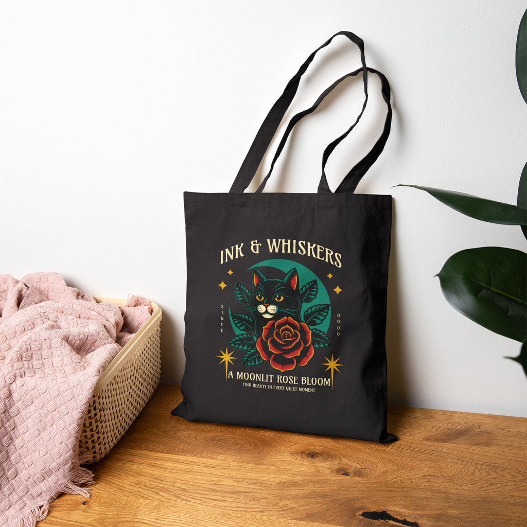 Black Ink & Whiskers tote bag with cat and rose design on a wooden surface with a white wall background