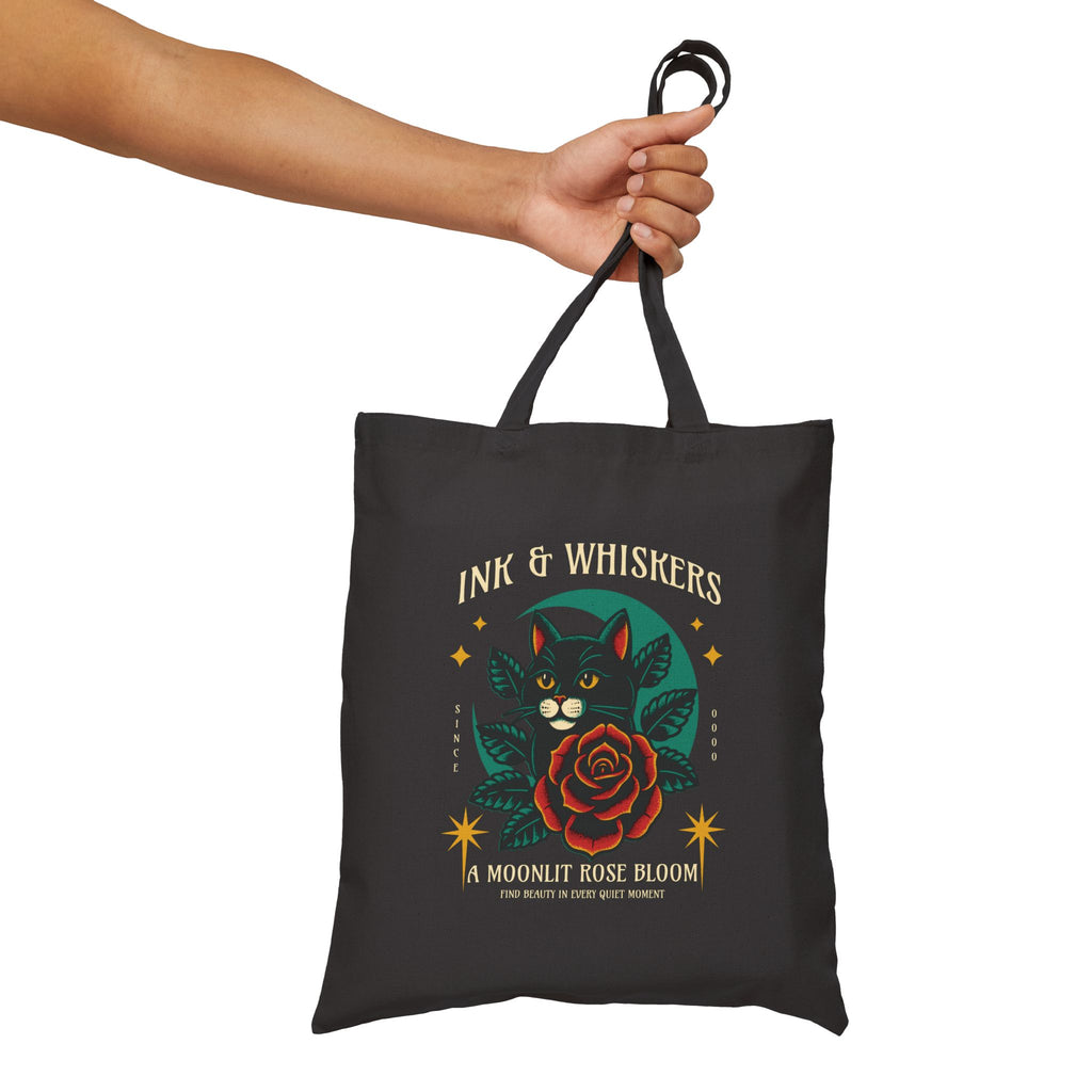 Black tote bag with cat and rose design held by a hand on a white background