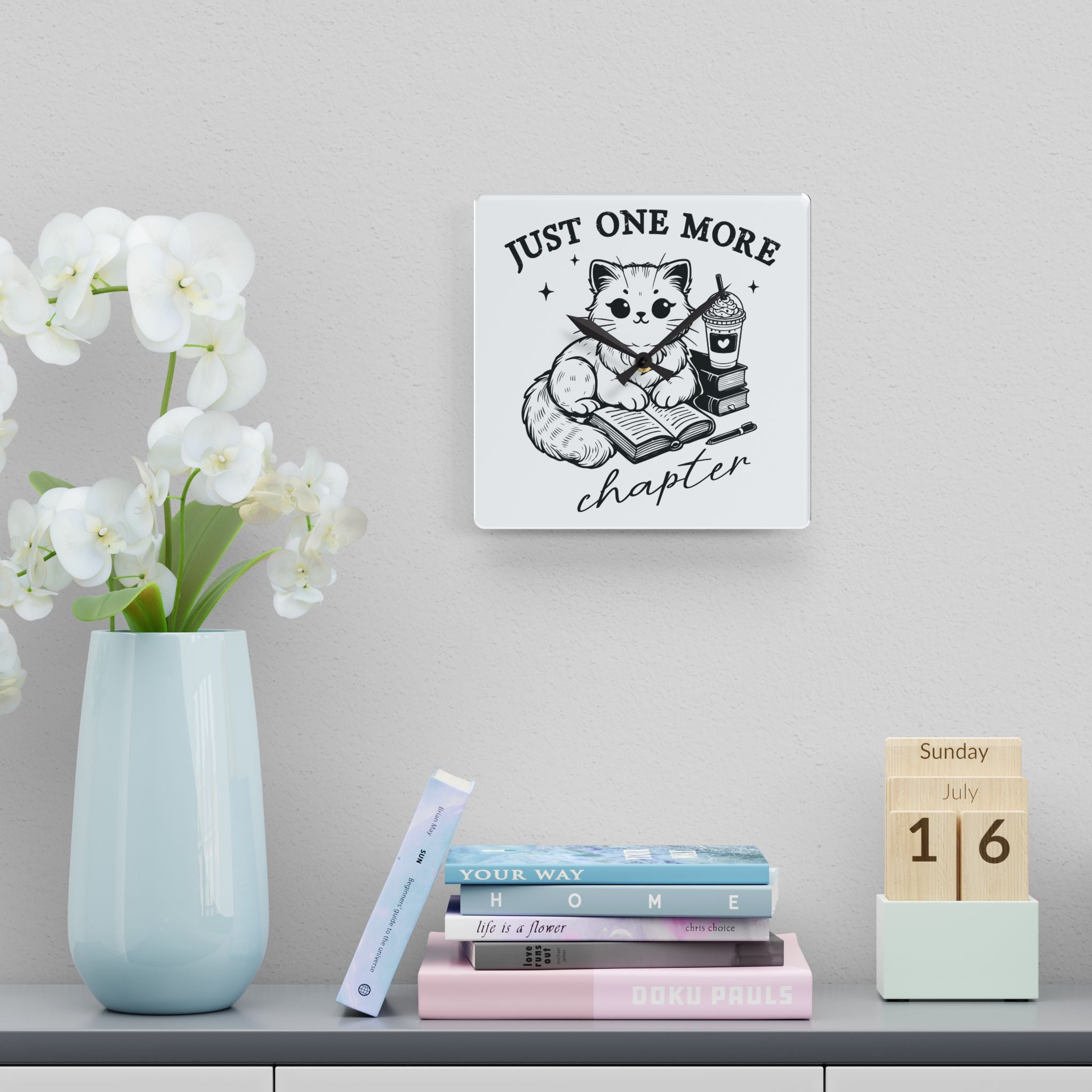Line art graphic on the "Just One More Chapter" Acrylic Wall Clock depicting a cat reading an open book beside a book stack topped with a beverage. The words "just one more" are above, and "chapter" in script is below.