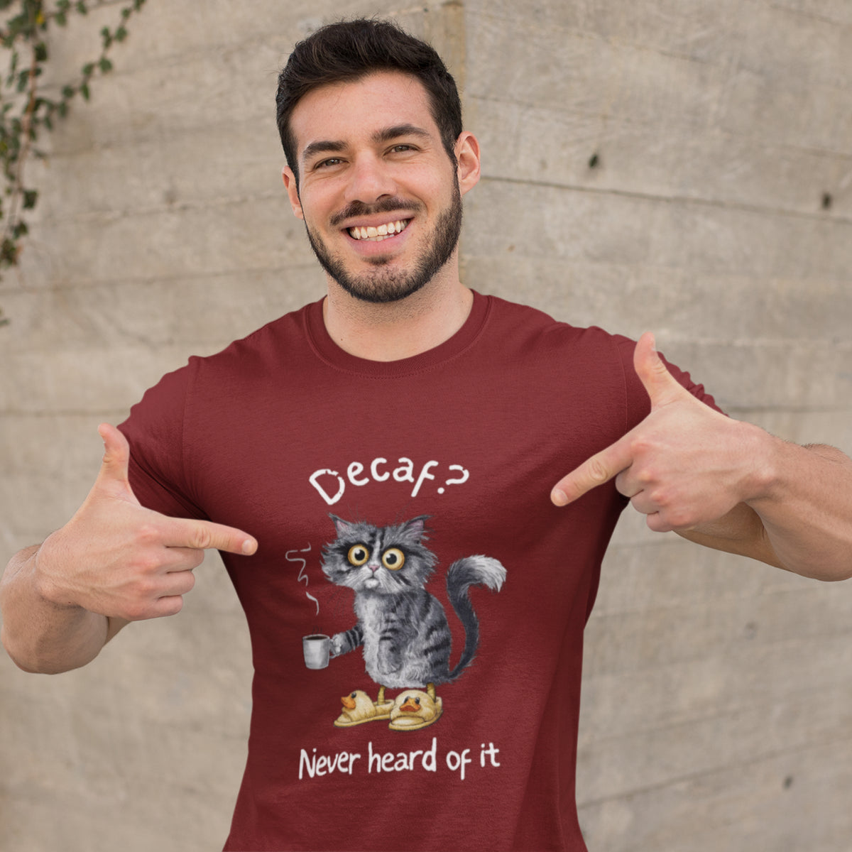 A man wearing a maroon t-shirt with a funny graphic of a gray cat holding a cup, with the text 'Decaf? Never heard of it'. He is pointing at the graphic with both thumbs, standing in front of a concrete wall.