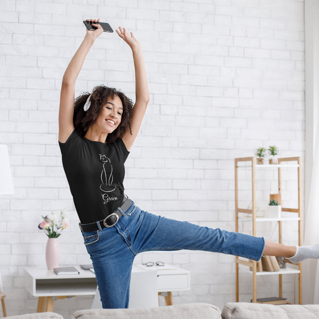 A woman in a black line art t-shirt and blue jeans poses dynamically in a bright, modern living room with a white brick wall background.