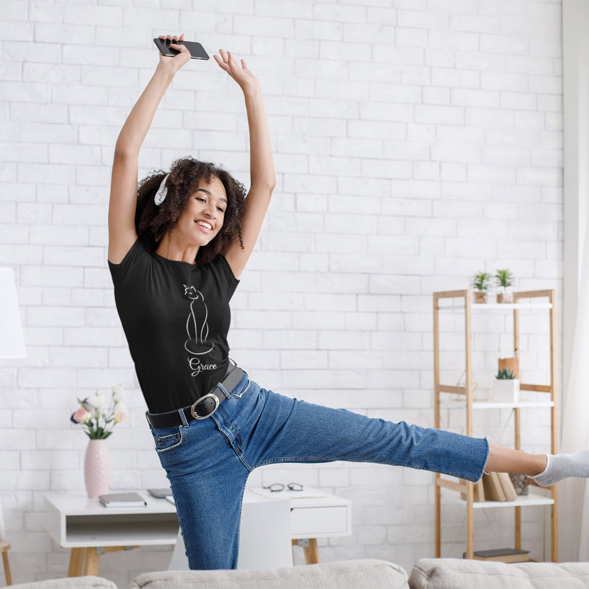 A woman in a black line art t-shirt and blue jeans poses dynamically in a bright, modern living room with a white brick wall background.