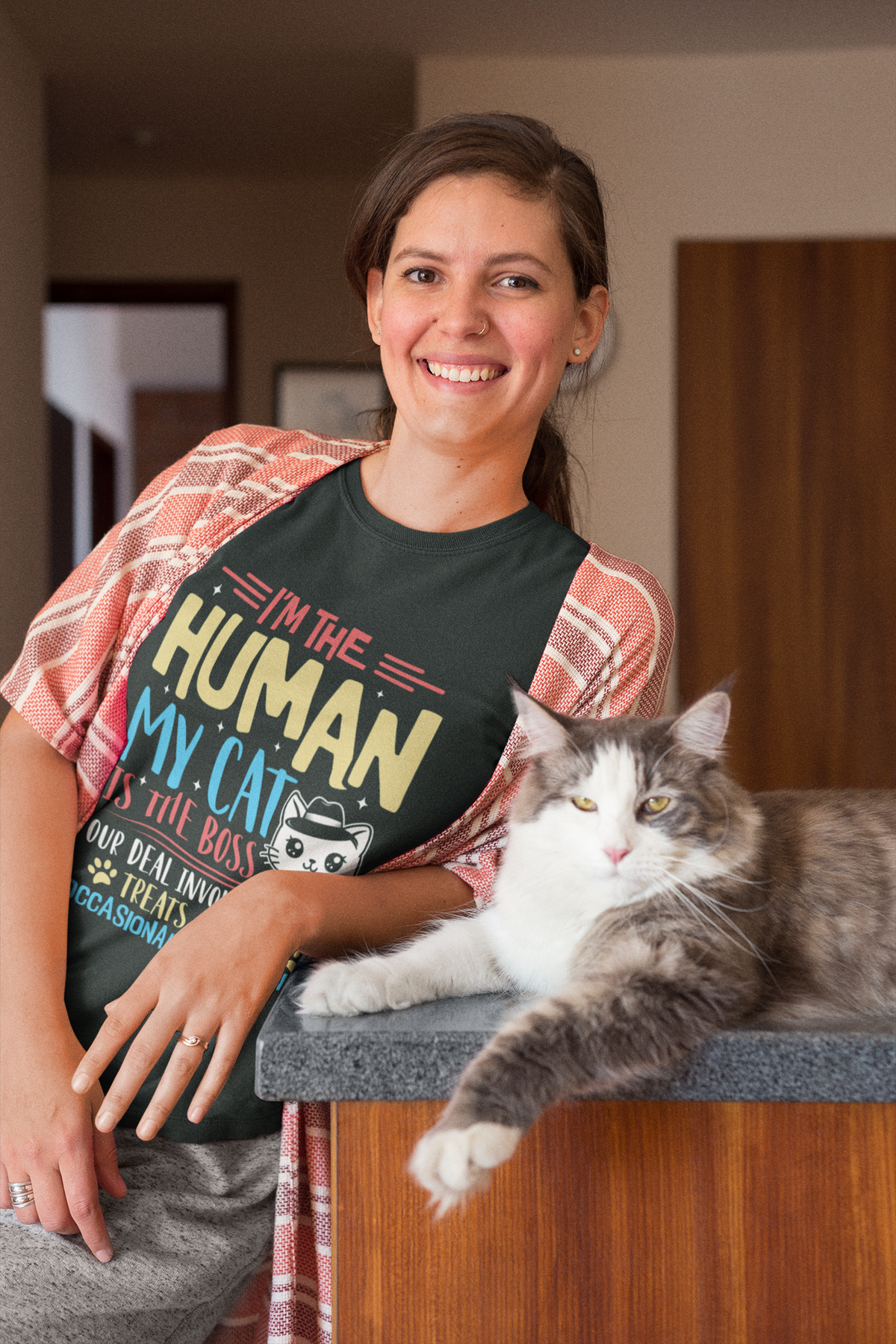 A woman smiling while wearing a graphic t-shirt that reads 'I'M THE HUMAN MY CAT IS THE BEST' with a cat illustration, with a fluffy gray and white cat resting on a kitchen counter next to her.