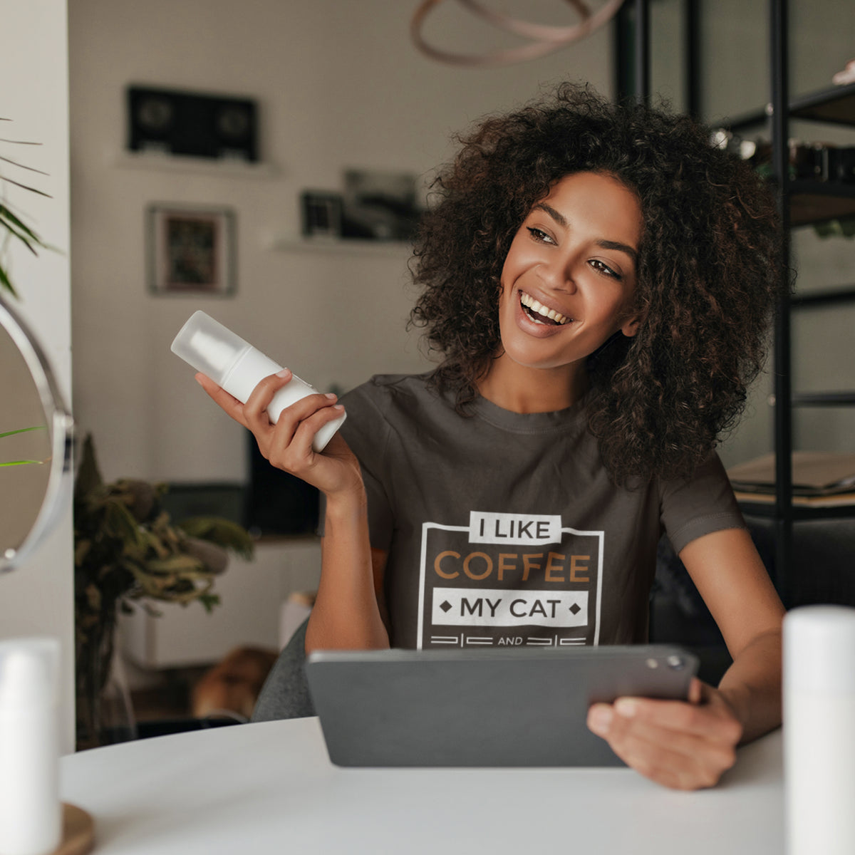 A woman with curly hair smiles while holding a white bottle in a home setting, wearing a brown t-shirt with the text 'I LIKE COFFEE MY CAT'. She is also holding a tablet on a white table.