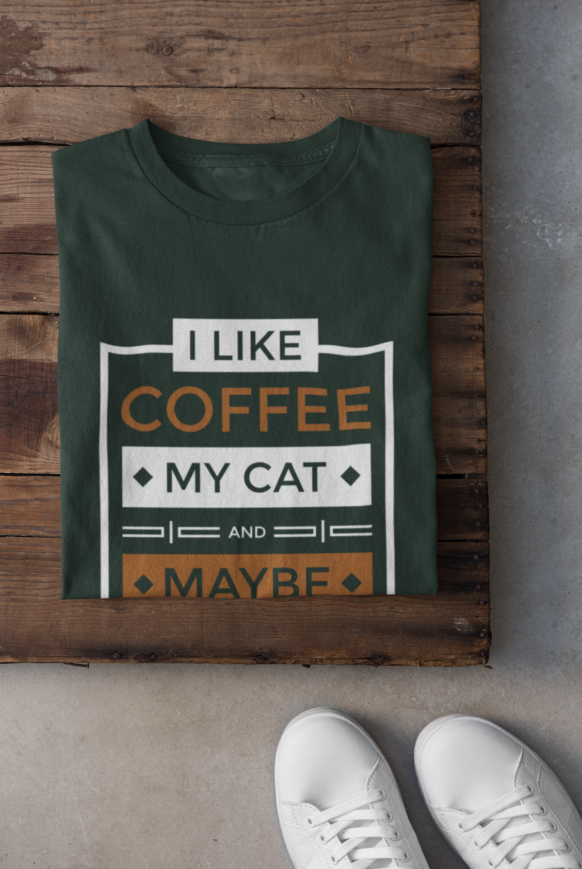 A dark green folded t-shirt with a graphic print reading 'I LIKE COFFEE MY CAT AND MAYBE' in white and orange, styled on a rustic wooden board with white sneakers visible at the bottom.