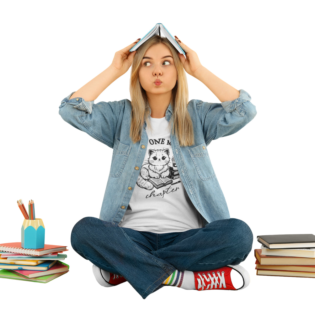 A young woman sits cross-legged on a white background, wearing a light blue denim shirt over a white graphic t-shirt, blue jeans, and red sneakers. She is holding an open book above her head like a hat, with books and a pencil cup on either side of her.