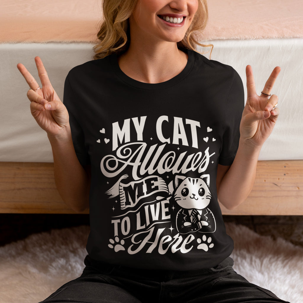 A woman wearing a black graphic t-shirt with a white print that reads 'MY CAT Allows ME TO LIVE Here' with a cartoon cat in a suit. She is sitting in front of a bed, making peace signs with both hands.