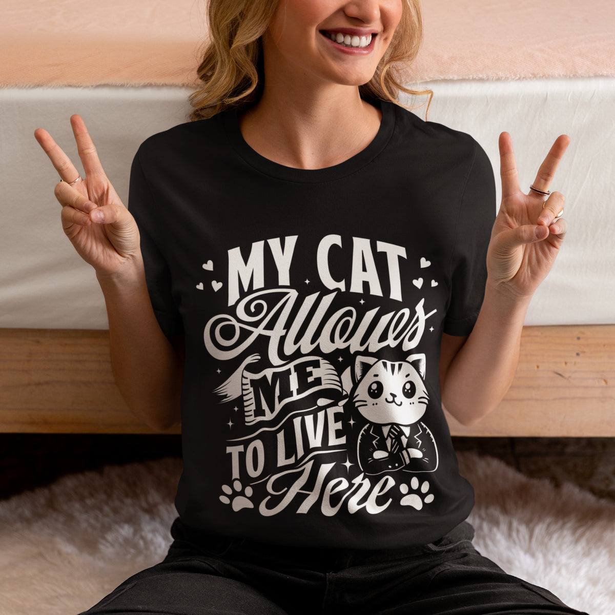 A woman wearing a black graphic t-shirt with a white print that reads 'MY CAT Allows ME TO LIVE Here' with a cartoon cat in a suit. She is sitting in front of a bed, making peace signs with both hands.