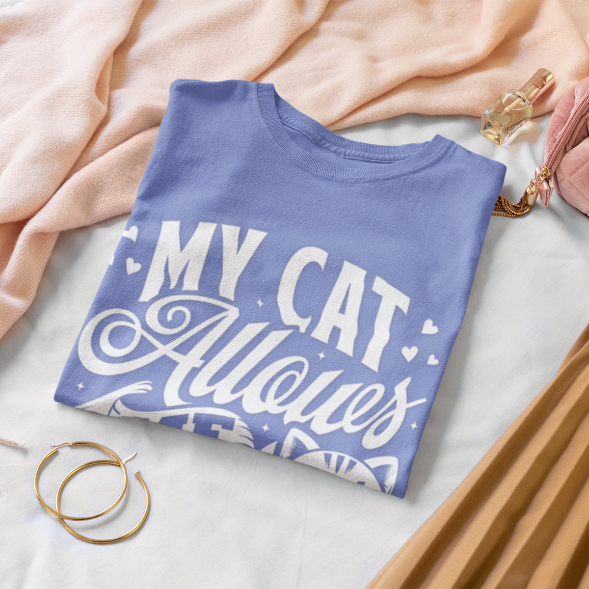 A folded periwinkle blue t-shirt with a white graphic reading 'MY CAT Allows Me' is styled in a flat-lay arrangement on a white sheet with a peach blanket, gold hoop earrings, a perfume bottle, and a pink clutch.