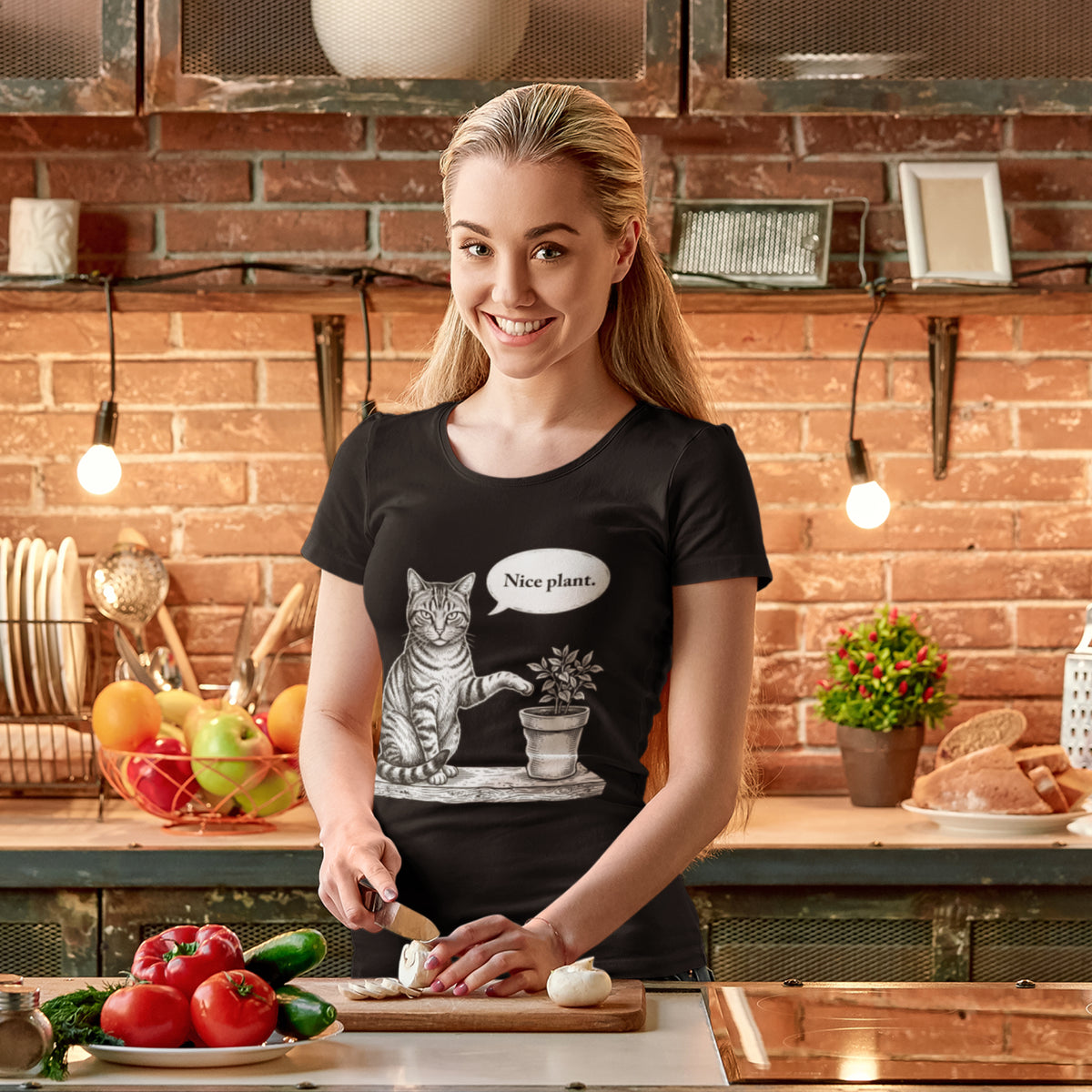 A woman in a kitchen wearing a black graphic t-shirt with a cat sitting next to a potted plant and a speech bubble that says 'Nice plant.' She is cutting vegetables on a wooden board in front of a brick wall background.