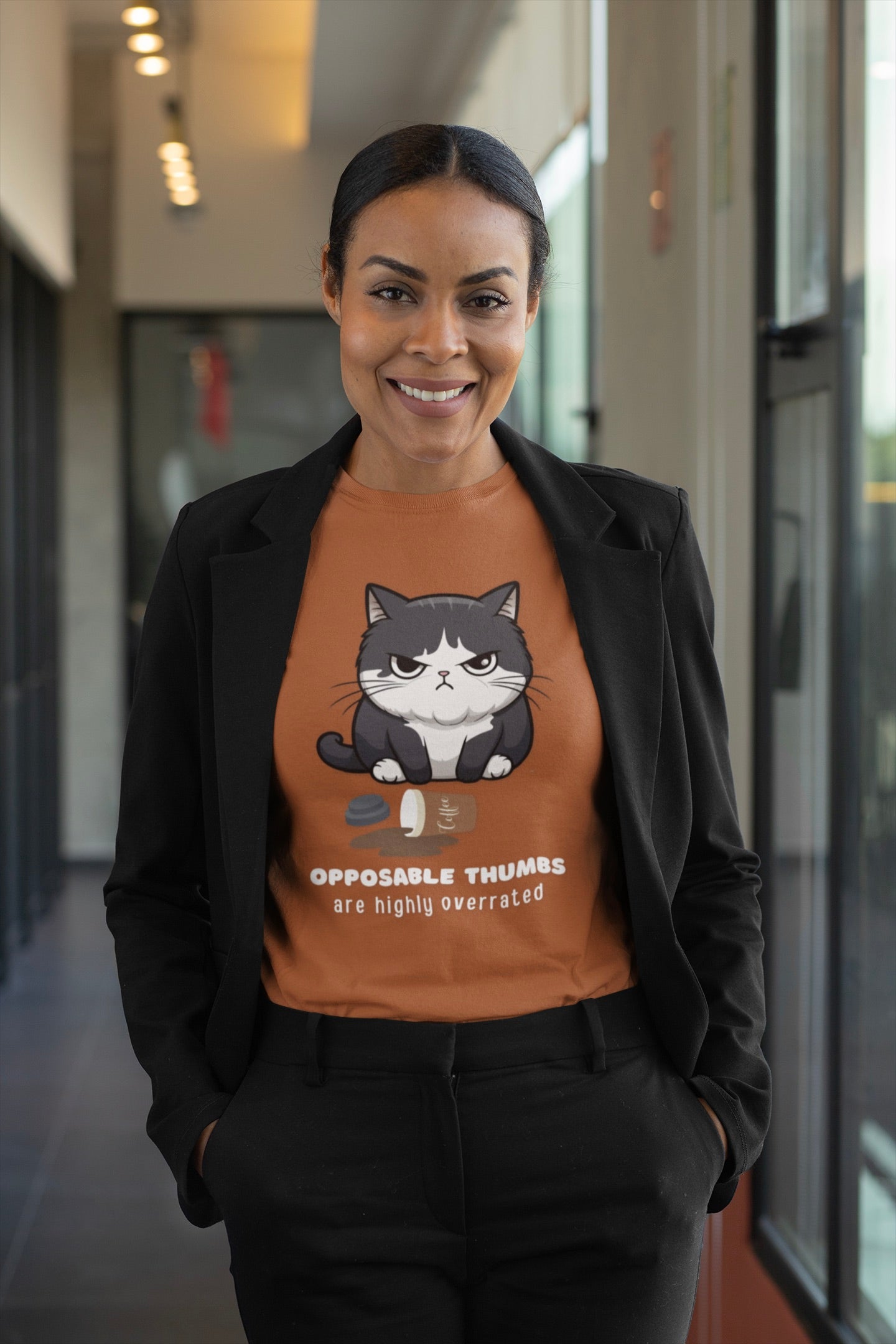A woman wearing a black blazer and black trousers stands in a hallway, wearing an orange crewneck t-shirt with a cartoon cat graphic and the text 'OPPOSABLE THUMBS are highly overrated'.