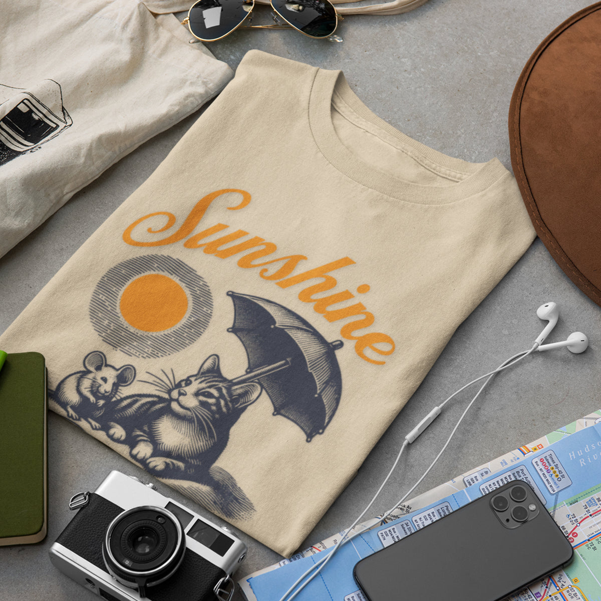 A beige graphic T-shirt laid flat on a gray surface, surrounded by styled props including sunglasses, earbuds, a camera, a phone, a notebook, and a hat. The shirt features the word 'Sunshine' in orange script above a cat and mouse illustration holding a dark umbrella, with a circular sun graphic.
