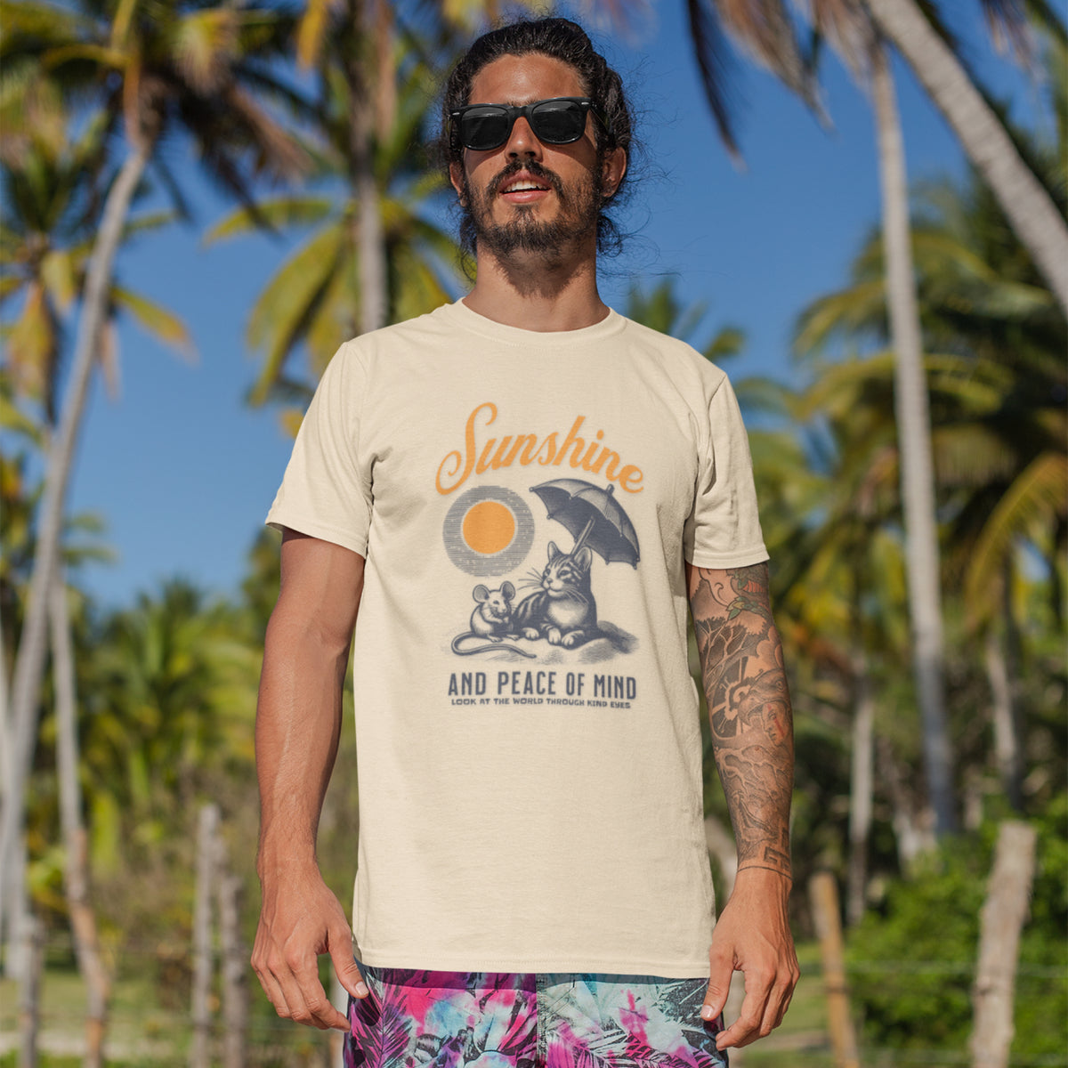 A man wearing a cream-colored graphic t-shirt with a cat and mouse sun design, standing outdoors in front of a tropical background of palm trees and blue sky.
