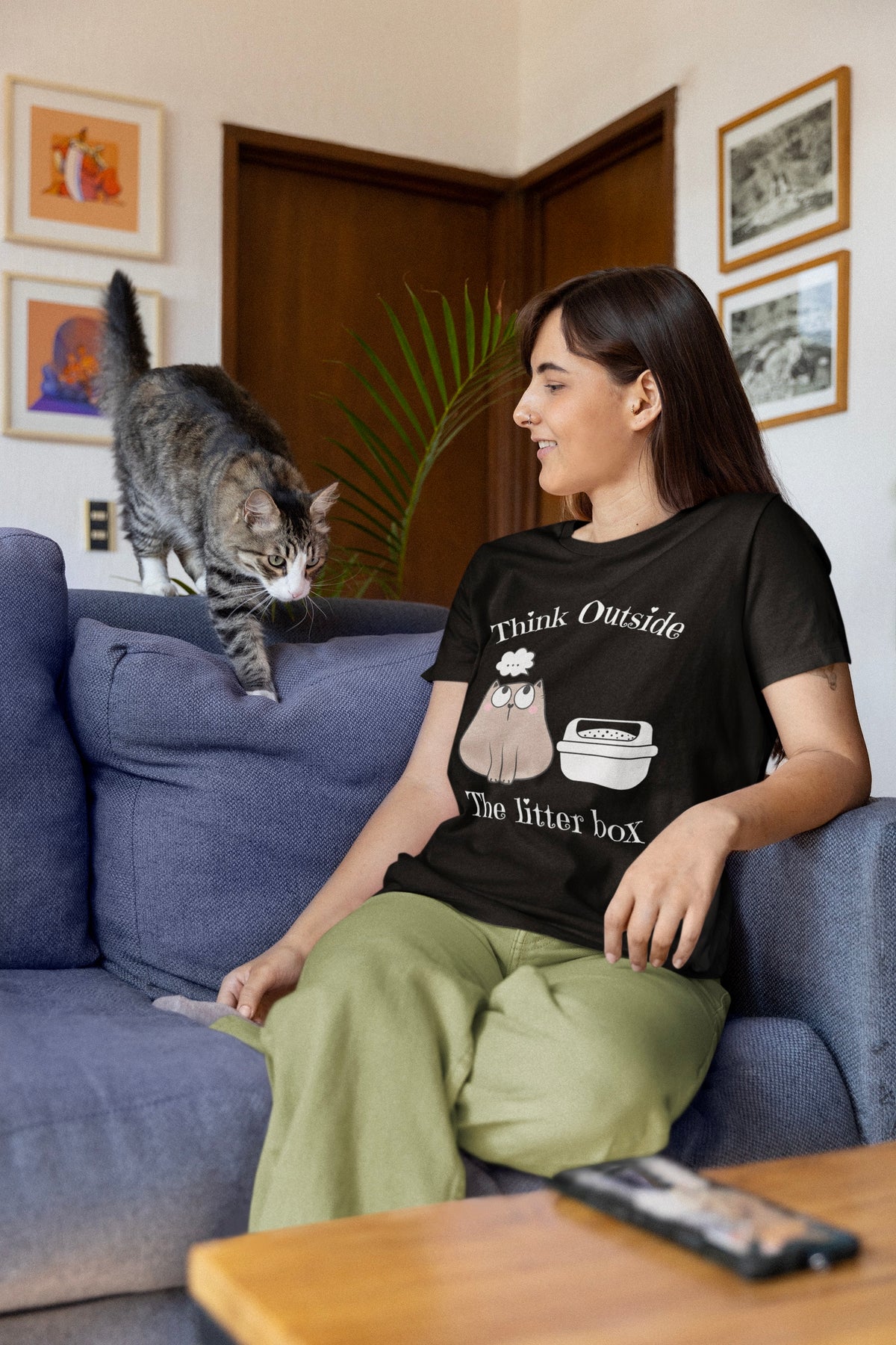 A woman sitting on a blue sofa in a living room, wearing a black graphic t-shirt that reads 'Think Outside The Litter box' with a graphic of a litter box. A gray tabby cat is walking along the back of the sofa beside her.