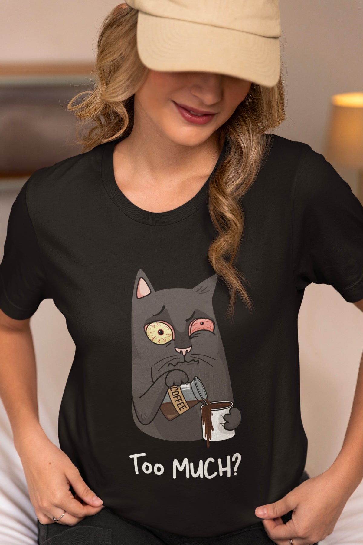 A woman wearing a black graphic t-shirt with a cartoon cat holding a coffee mug and the text 'Too Much?' below it, paired with a beige baseball cap.