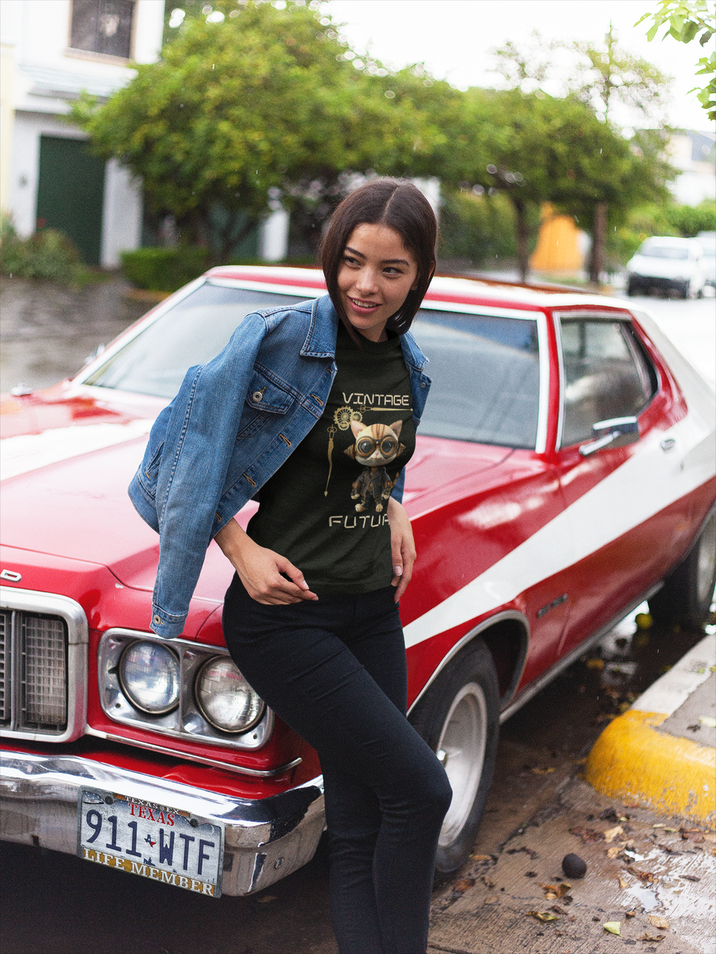 A woman posing in front of a red classic muscle car, wearing a black graphic t-shirt with a cat design and the words 'VINTAGE FUTURE', a blue denim jacket, and black pants.