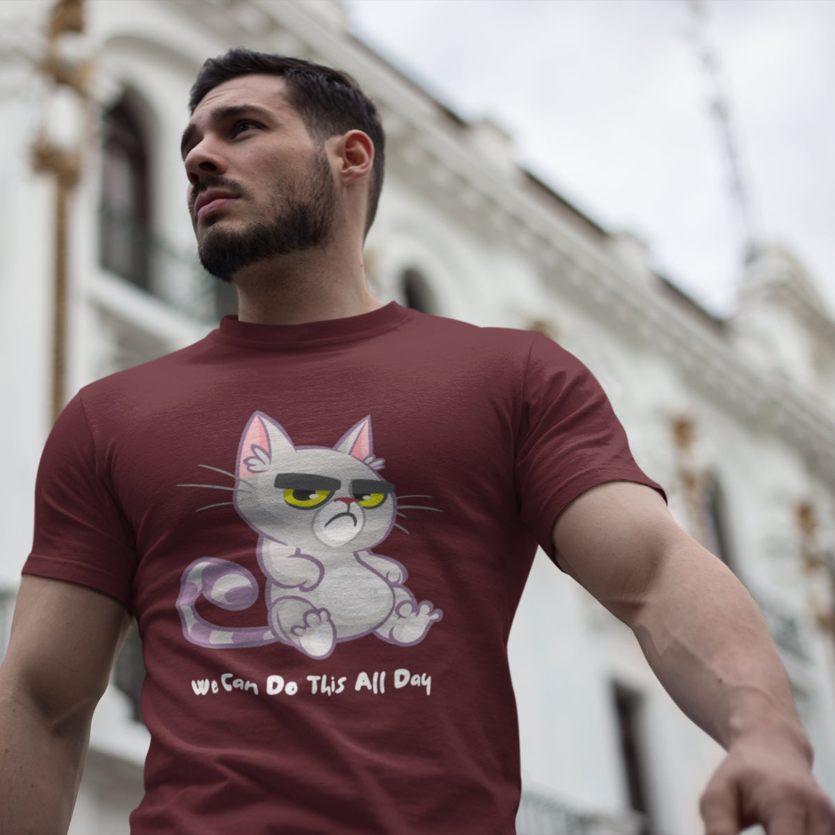A man wearing a maroon t-shirt with a cartoon graphic of a grumpy white cat  and the text 'WE CAN DO THIS ALL DAY' underneath, set against a blurred background of a white building.
