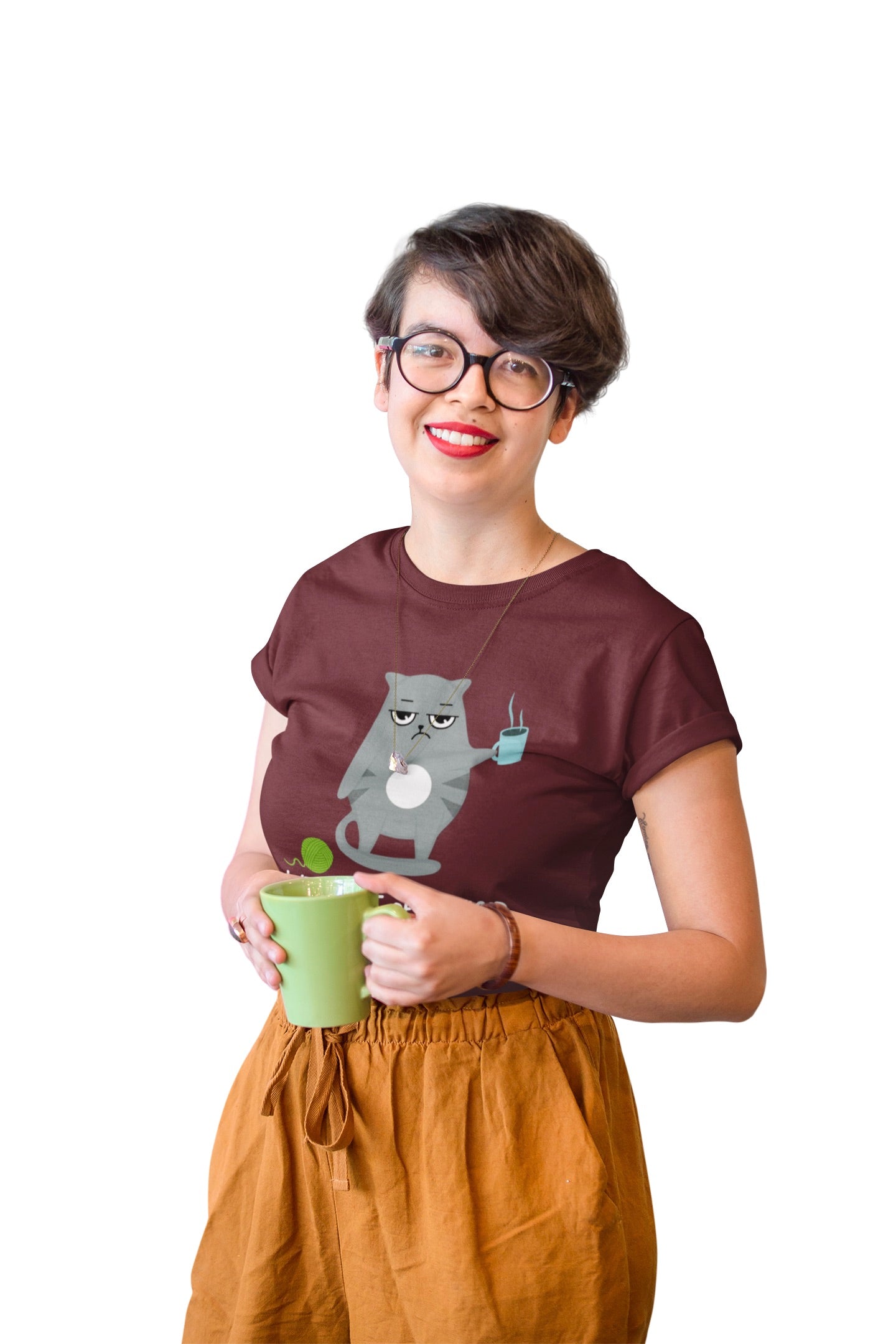 A woman with short brown hair and glasses wearing a dark brown t-shirt with a cartoon cat graphic and mustard yellow pants, holding a green mug against a plain white background.
