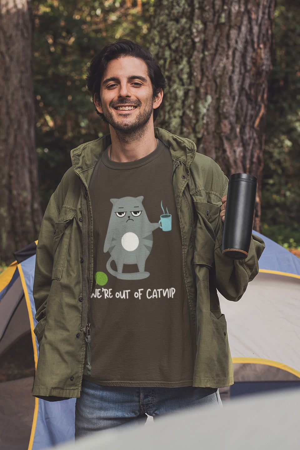 A man wearing a brown graphic t-shirt with a cartoon cat and the text 'WE'RE OUT OF CATMP' stands in front of a tent in a forest setting, holding a black travel mug.