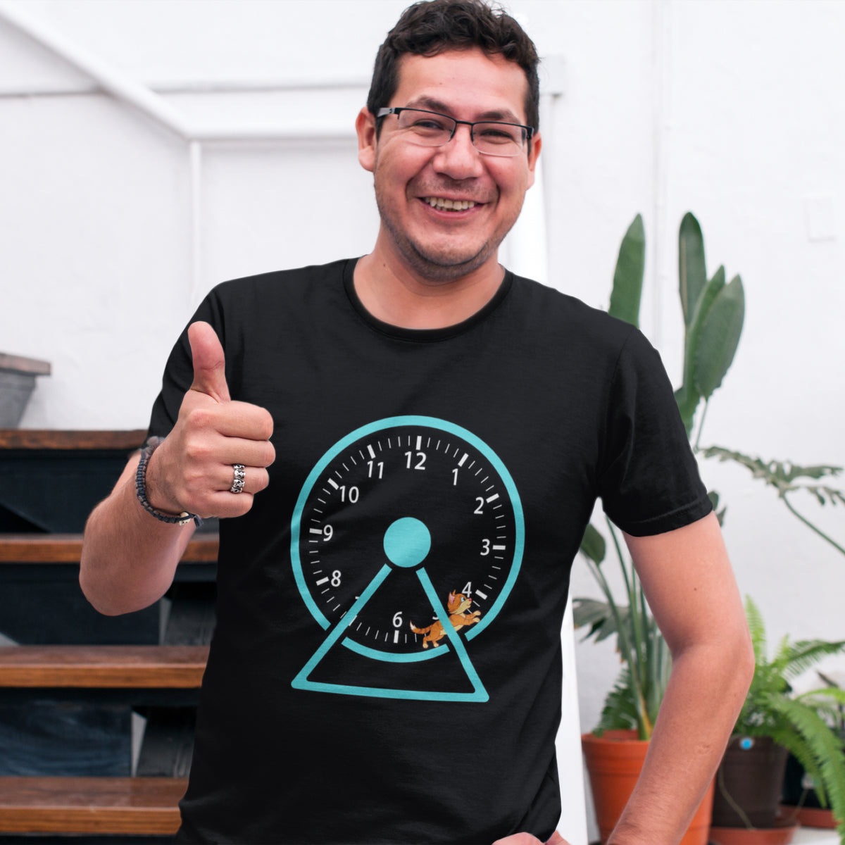 A smiling man wearing glasses and a black graphic t-shirt with a teal clock and triangle design, giving a thumbs-up in an indoor setting with plants and stairs in the background.