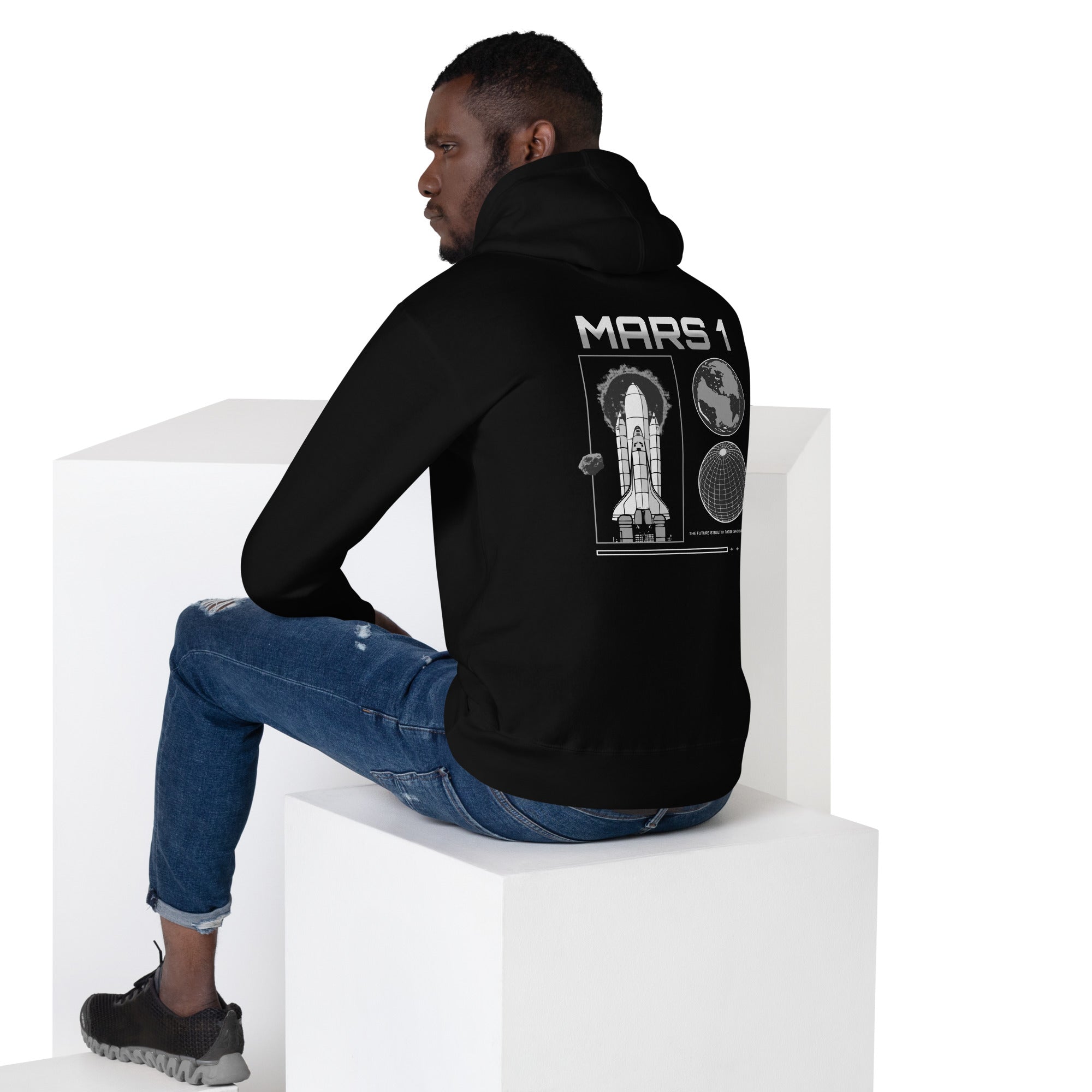  Model wearing a black hoodie with 'MARS 1' text above space shuttle and planet graphic on the back, sitting on a white cube.