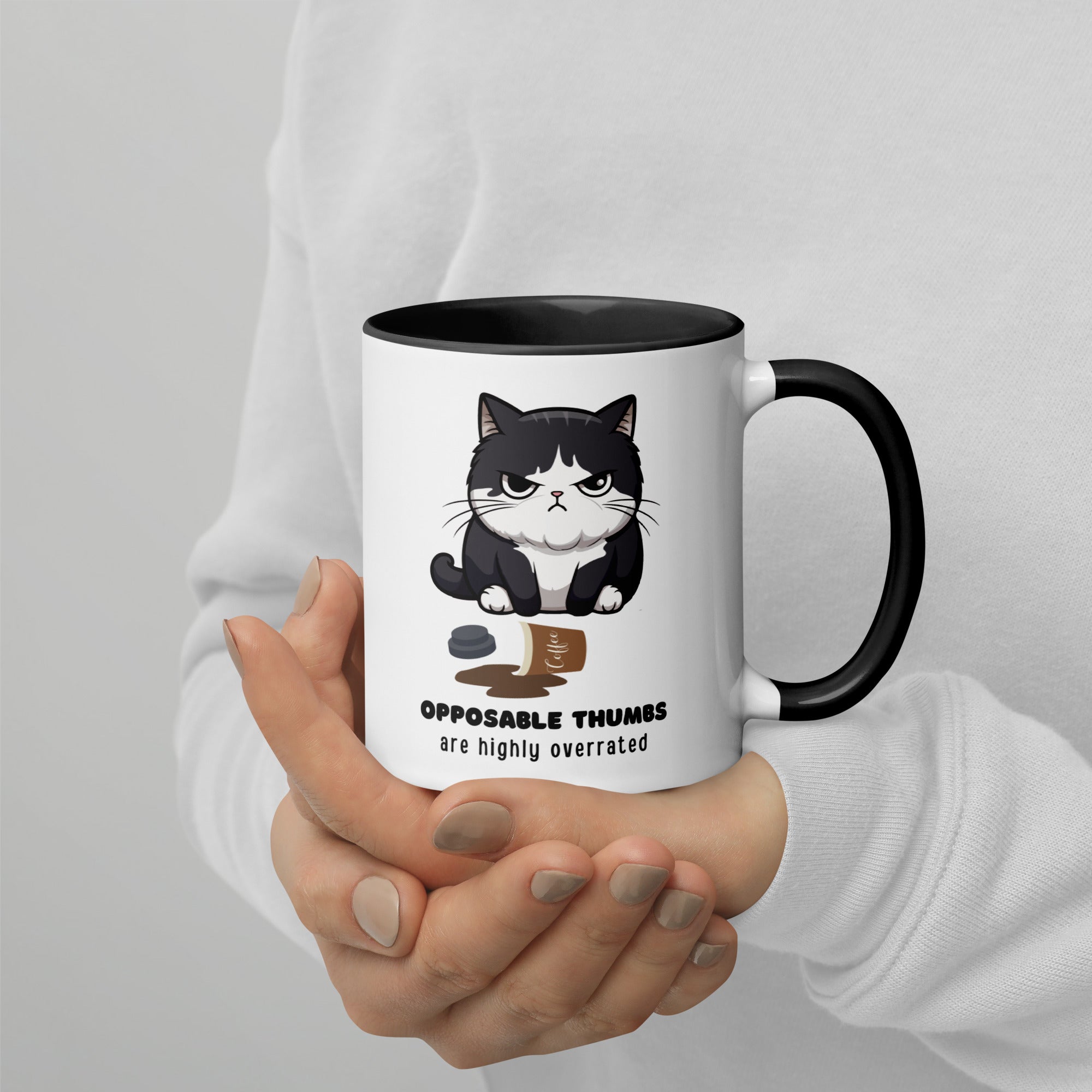 White and black ceramic mug with a graphic of a frustrated cartoon cat standing over spilled coffee. Text below the cat reads "Opposable Thumbs Are Highly Overrated".